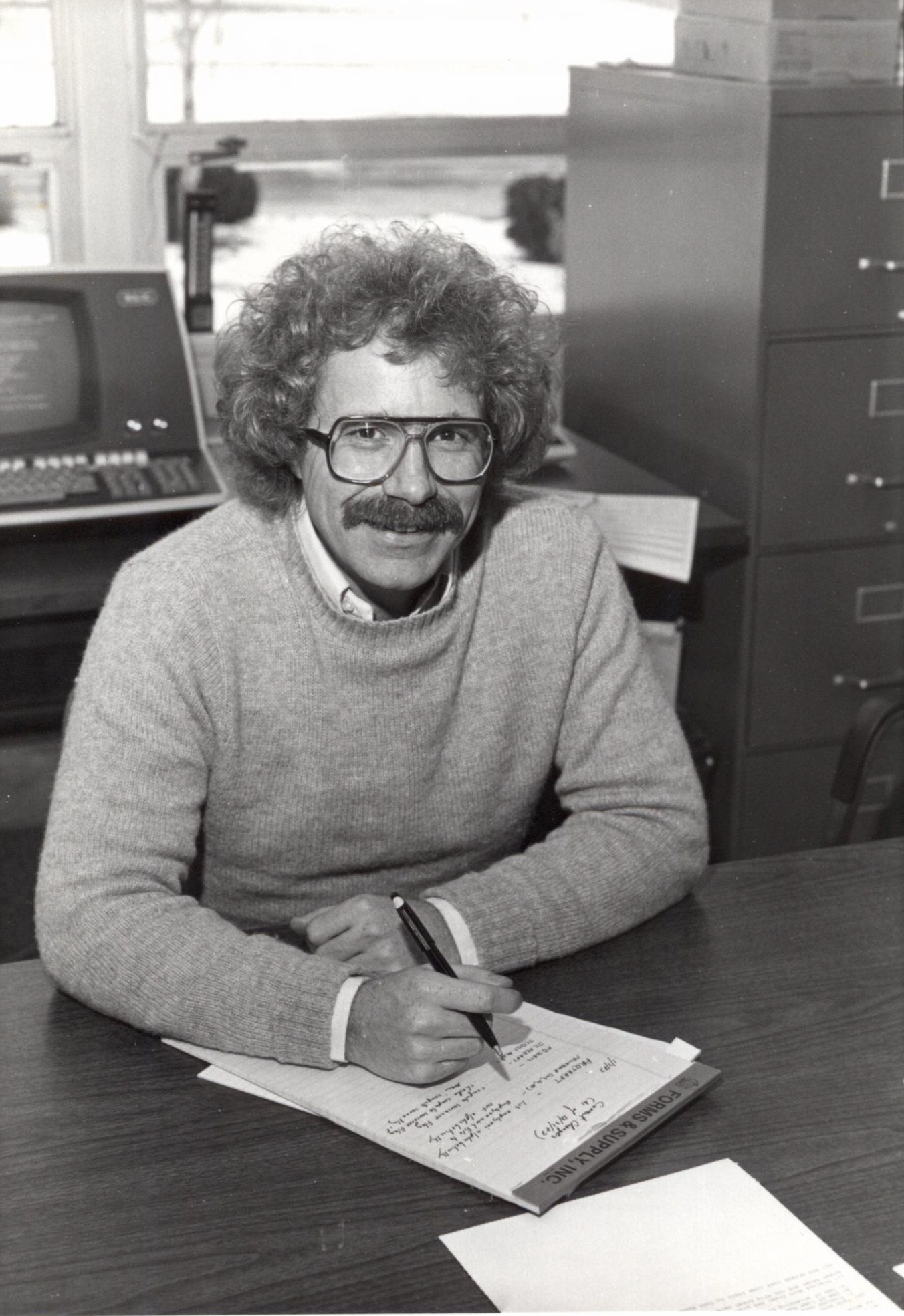 Fred Ode in the 1970s (black & white photo of a man taking notes on a legal pad at an office desk in front of an old computer - his hair is curly with a thick mustache, and he's wearing glasses)